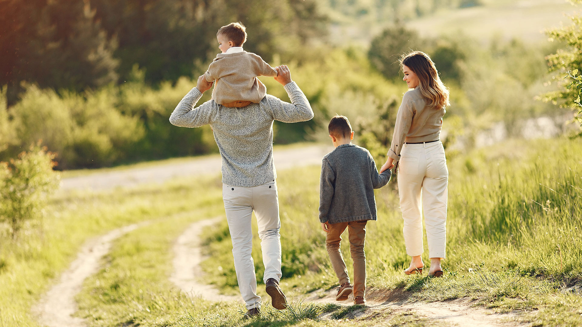 Seen from behind, a family walks down a grassy country path in warm sunlight, one son riding on his father's shoulders and another holding his mother's hand.