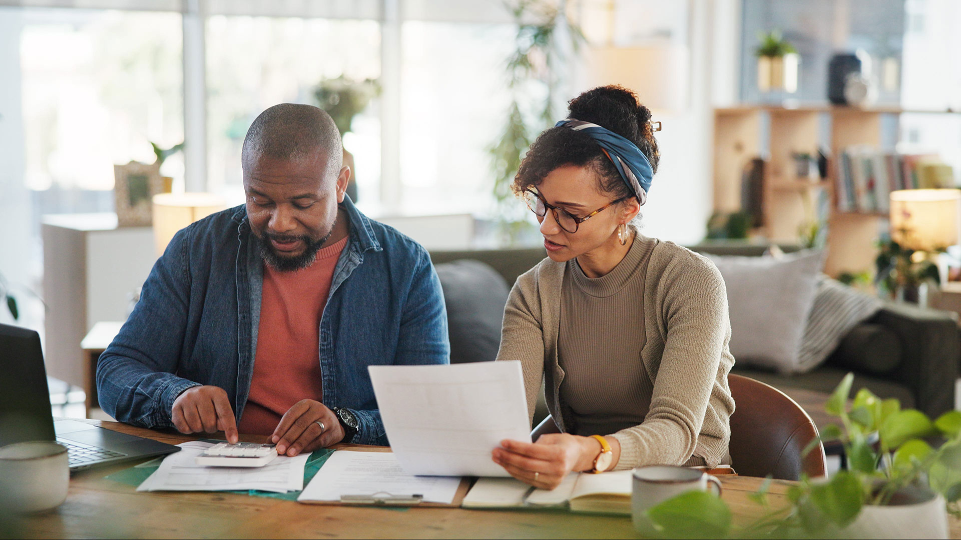 A man using a calculator and a woman holding a document review paperwork together at a table at home.