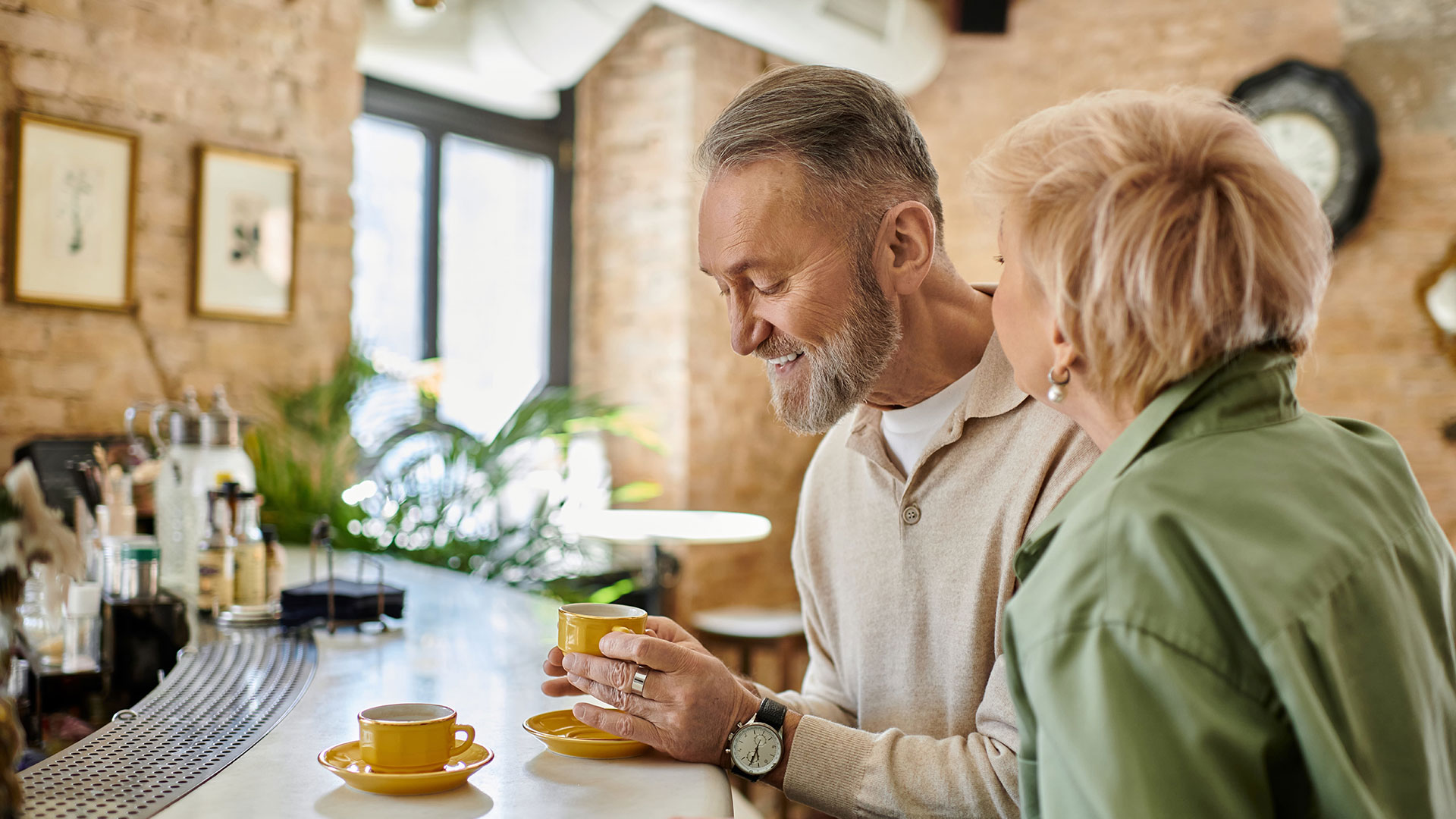 An older couple smile together over yellow cups of coffee at a café counter.