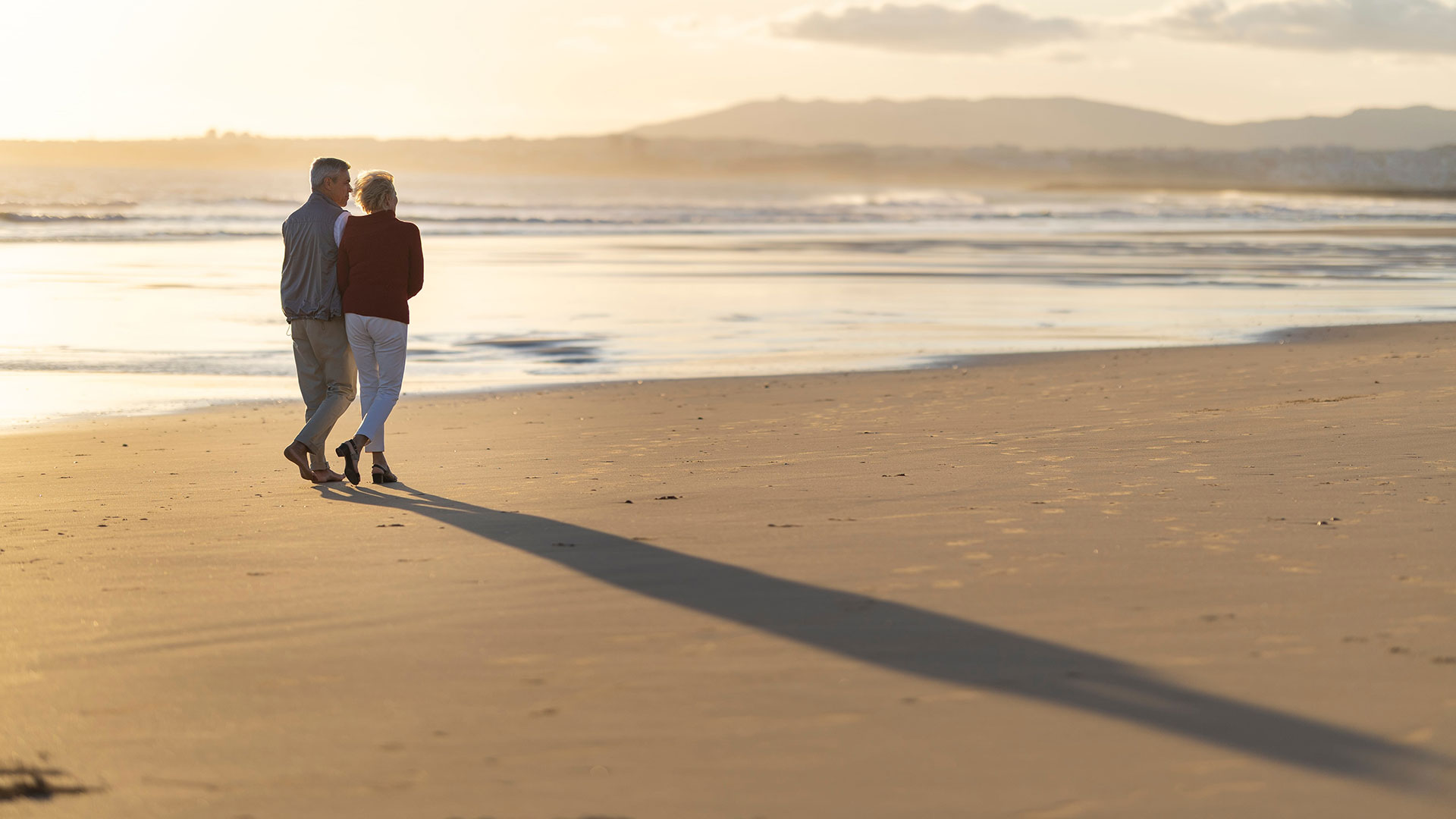 Seen from behind, an older couple walk arm in arm along a sandy beach at sunset, casting long shadows.