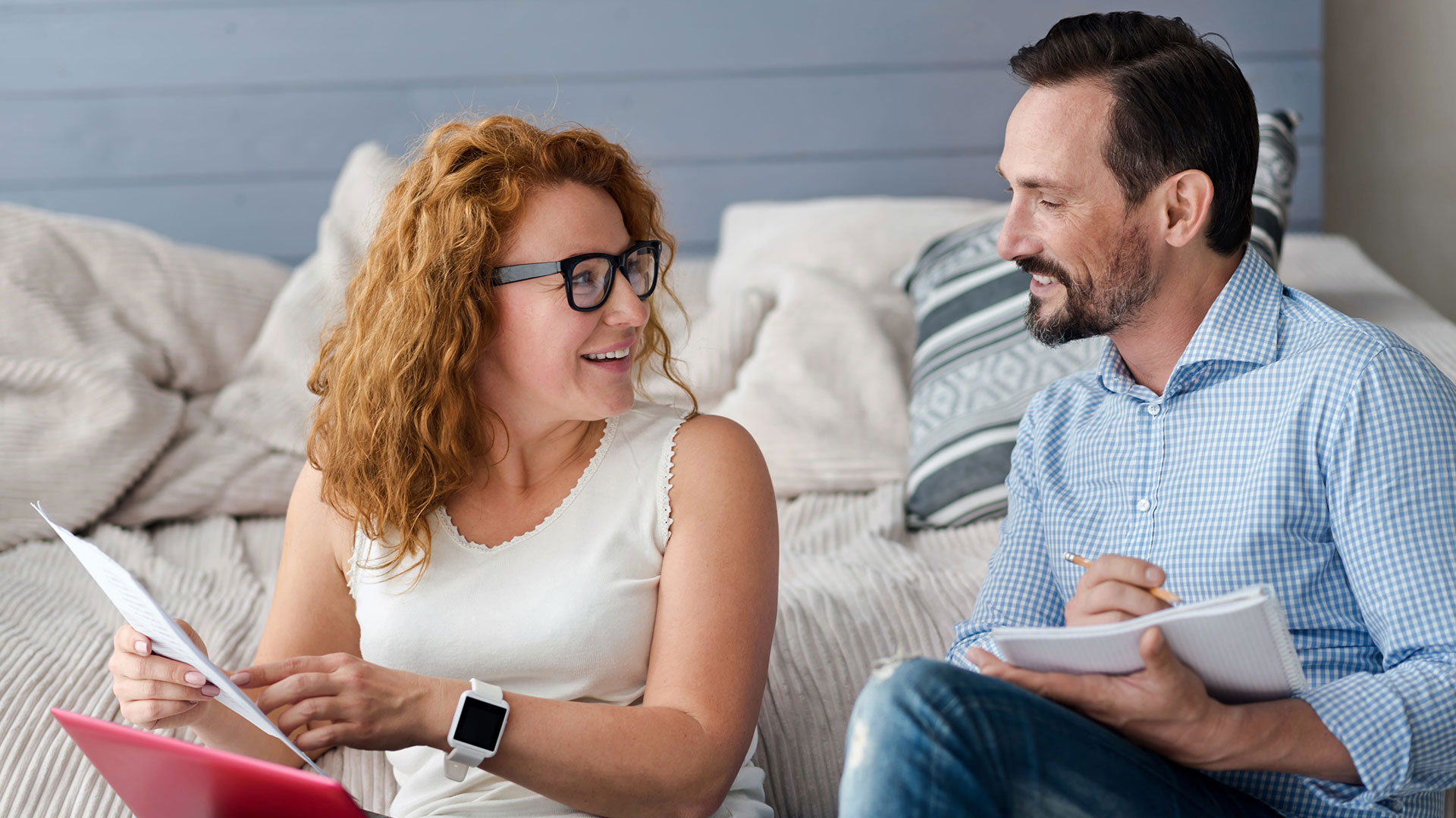 A red-haired woman in glasses holding a document smiles at a bearded man taking notes as they sit together on a bed.