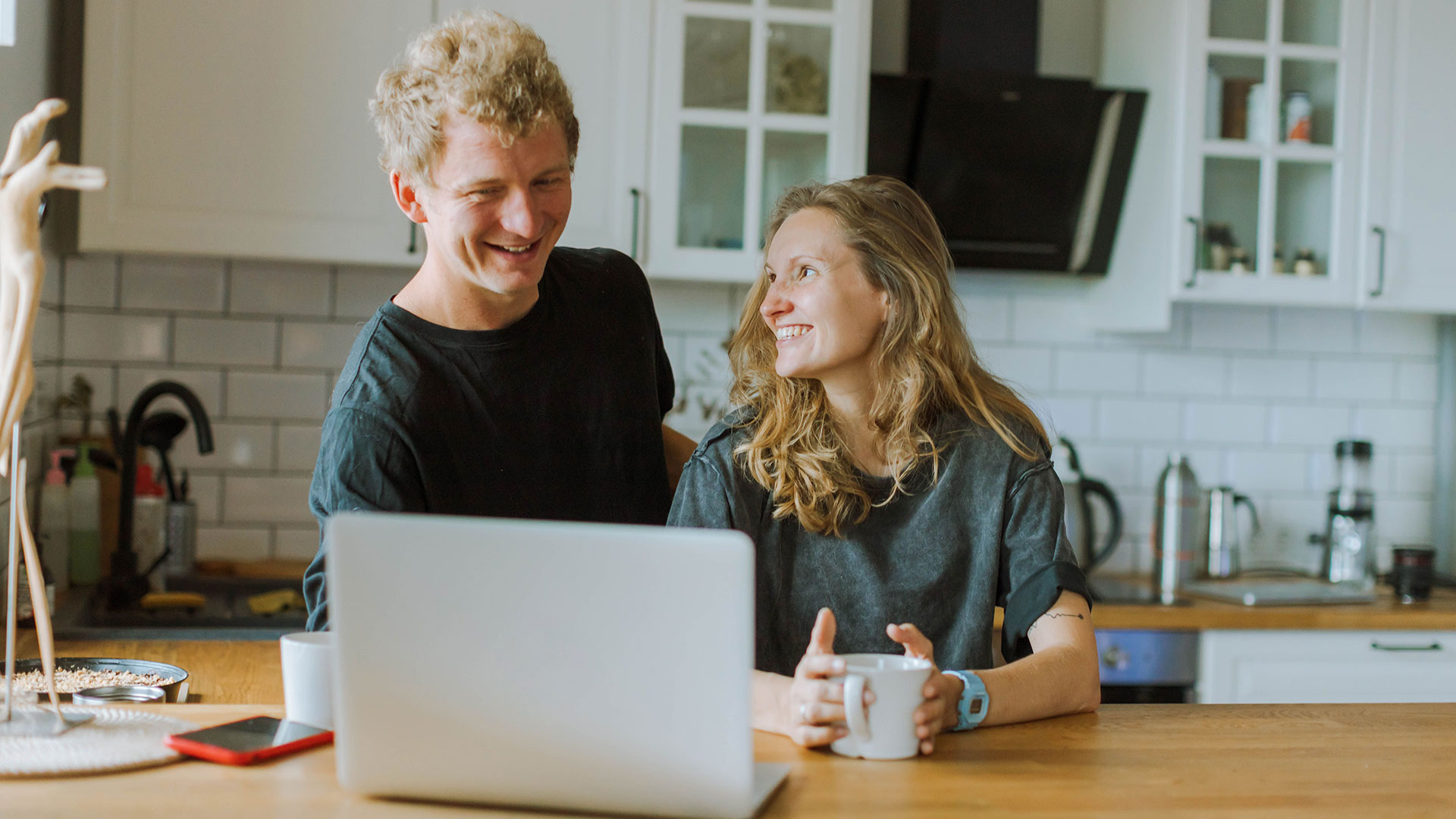 A blond man and a woman holding a mug smile at each other while sitting at a kitchen counter with a laptop.