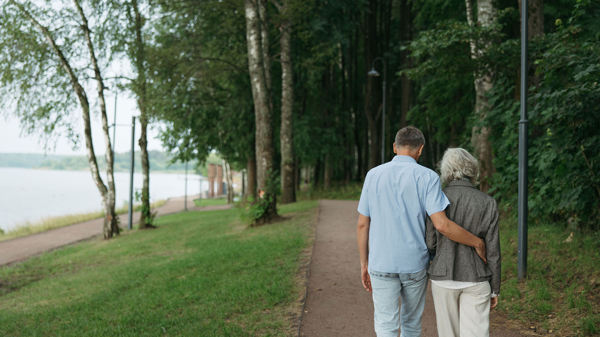 Seen from behind, an older couple walk arm in arm along a tree-lined path beside a lake.