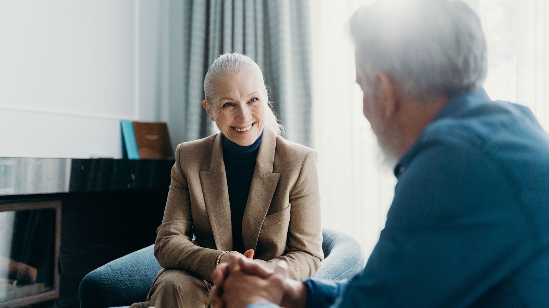 A silver-haired woman in a tan blazer smiles during a conversation with a man seated across from her by a window.