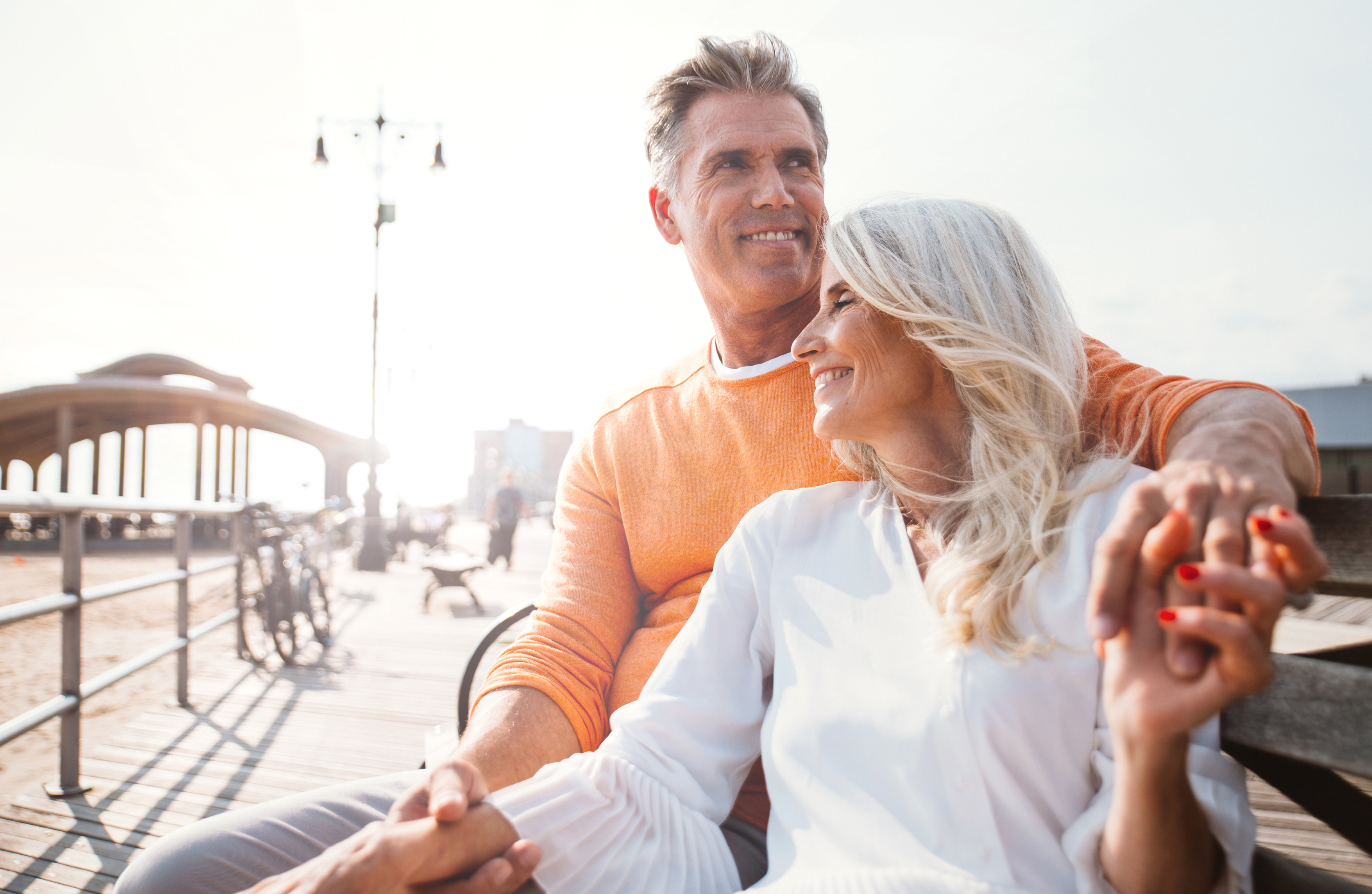 A smiling couple holds hands and enjoys the sunshine while sitting on a boardwalk bench