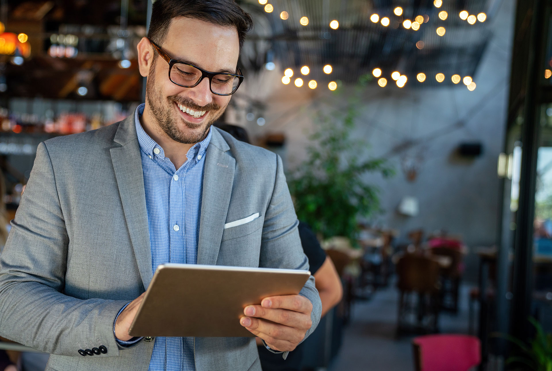 A man in a suit smiles while looking at a tablet inside a modern, warmly lit café