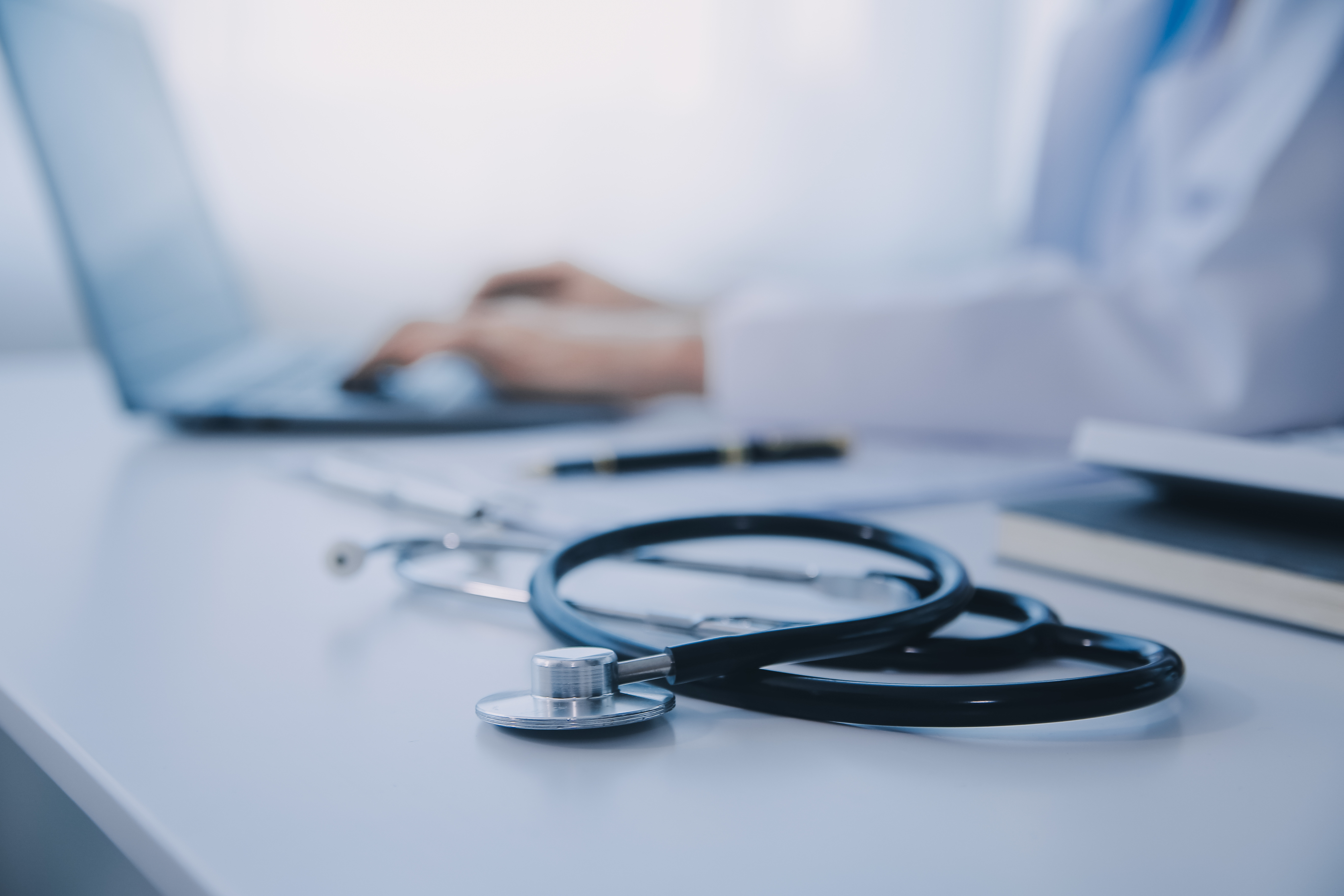 A stethoscope rests on a desk beside a laptop and notebook in a bright medical workspace