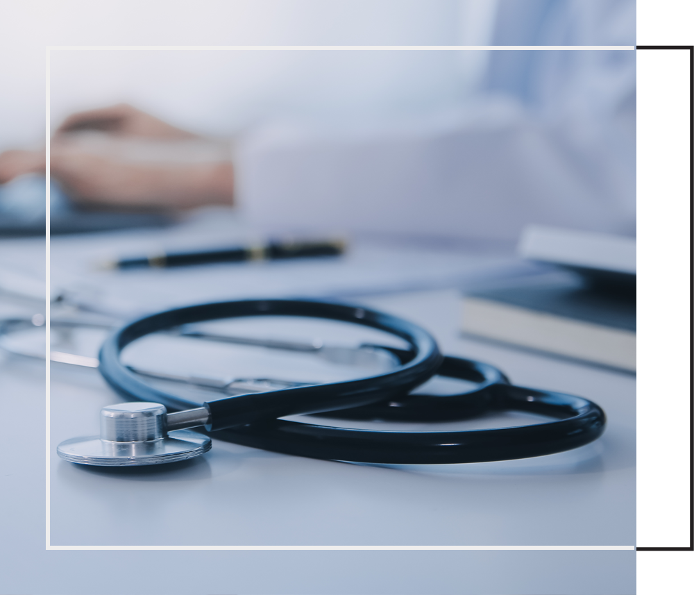 A stethoscope rests on a desk beside a laptop and notebook in a medical office setting.
