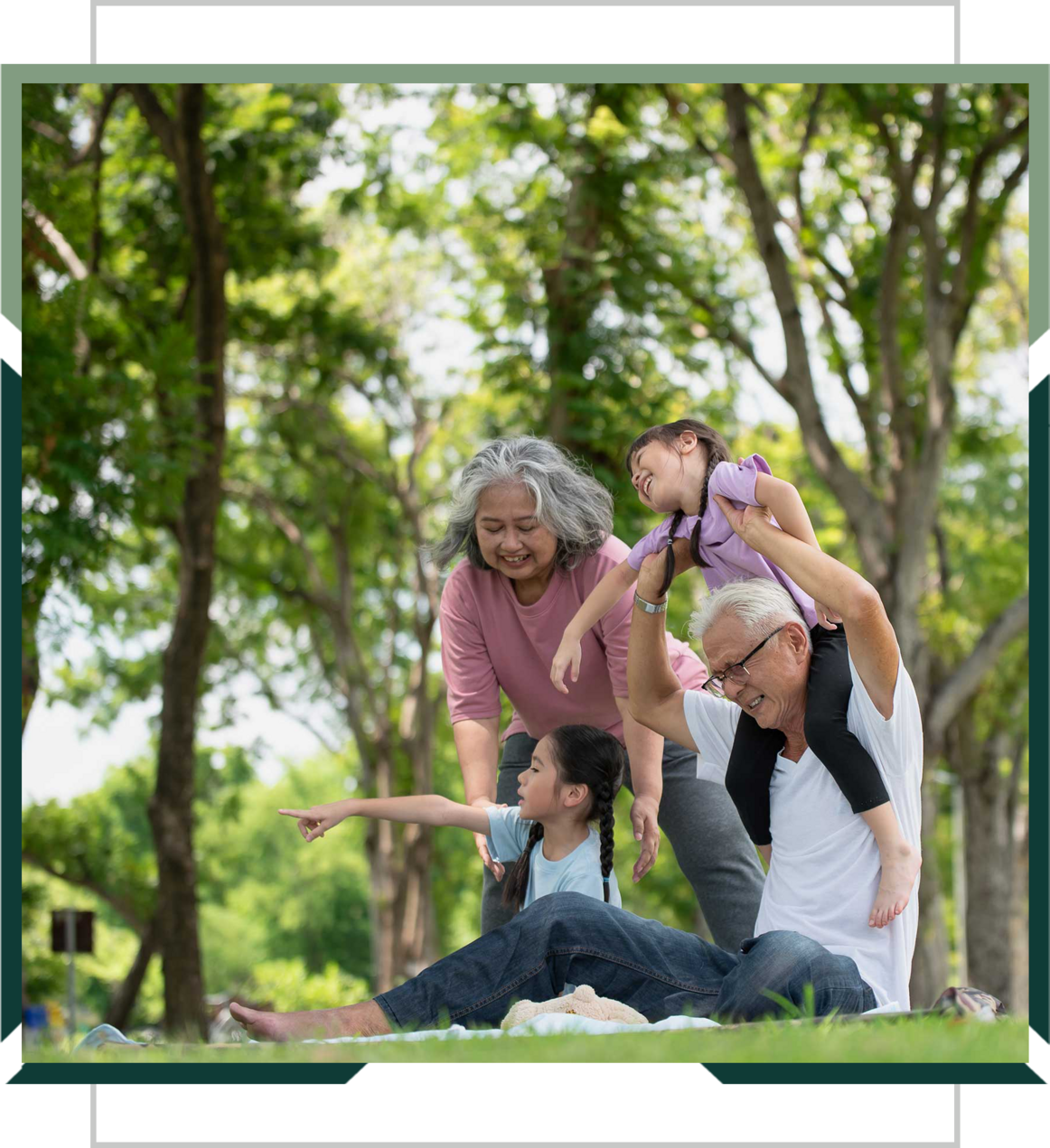 Two older adults playing with two young girls on a blanket in a green park.