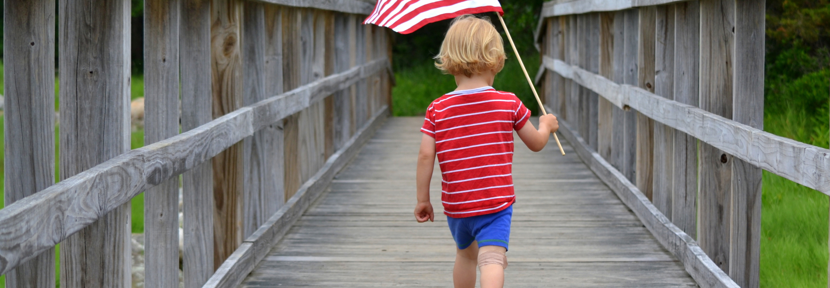 Young boy carrying an American Flag over a wooden bridge ​