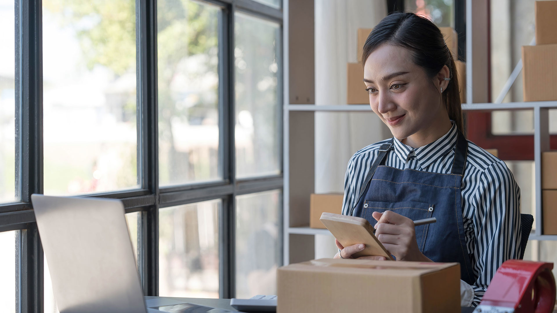 A woman wearing an apron writing on a notepad next to a laptop and a shipping box at a desk.
