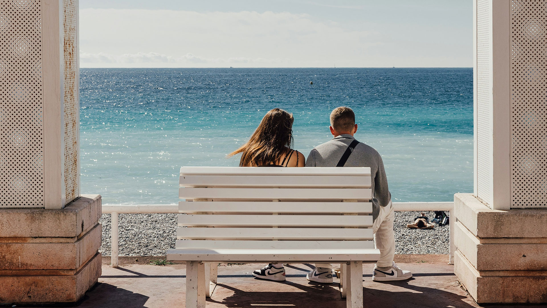 Two people sitting on a white bench looking out at the ocean from a covered walkway.