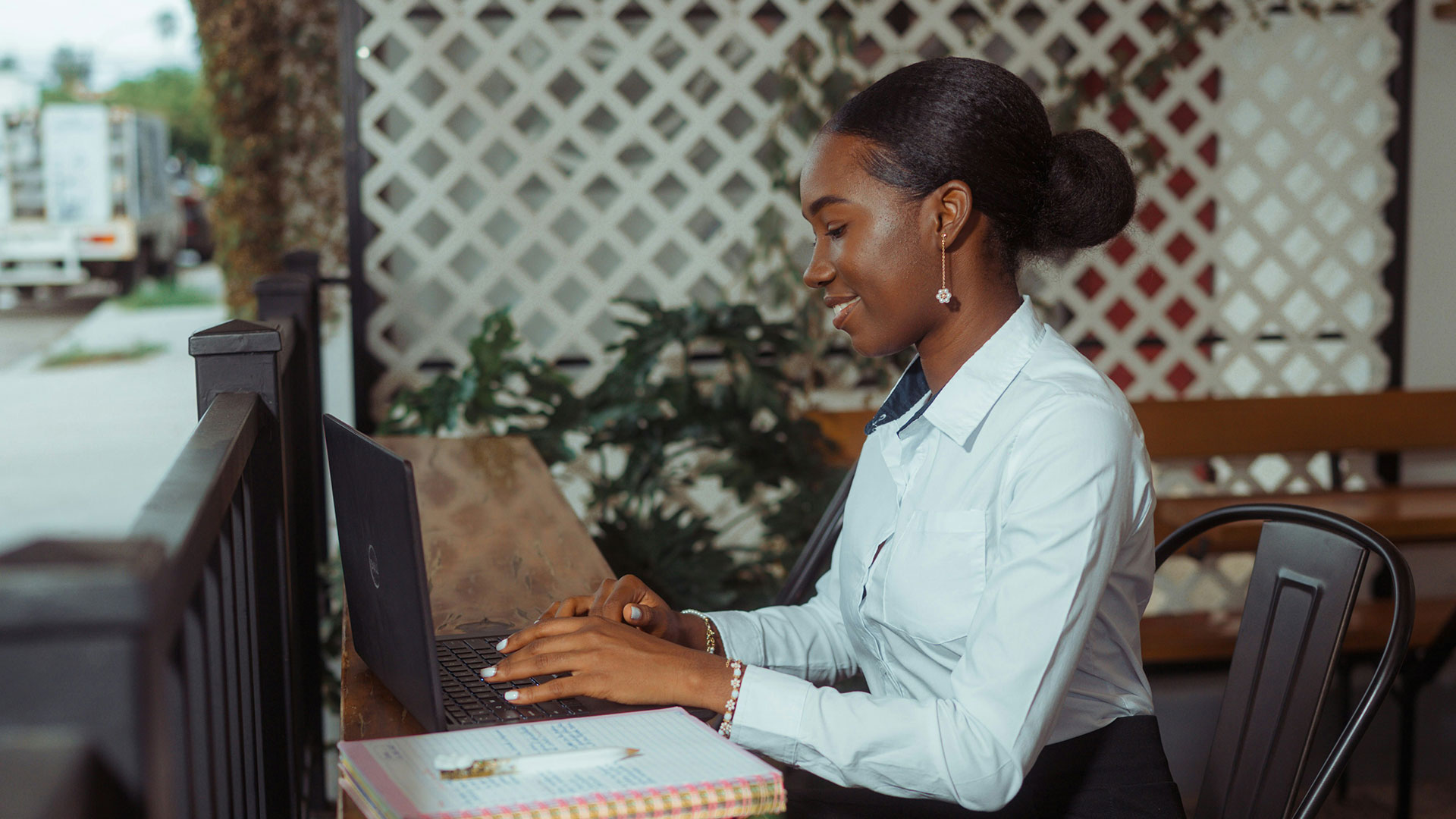 A woman working on a laptop at an outdoor cafe table with a notebook beside her.