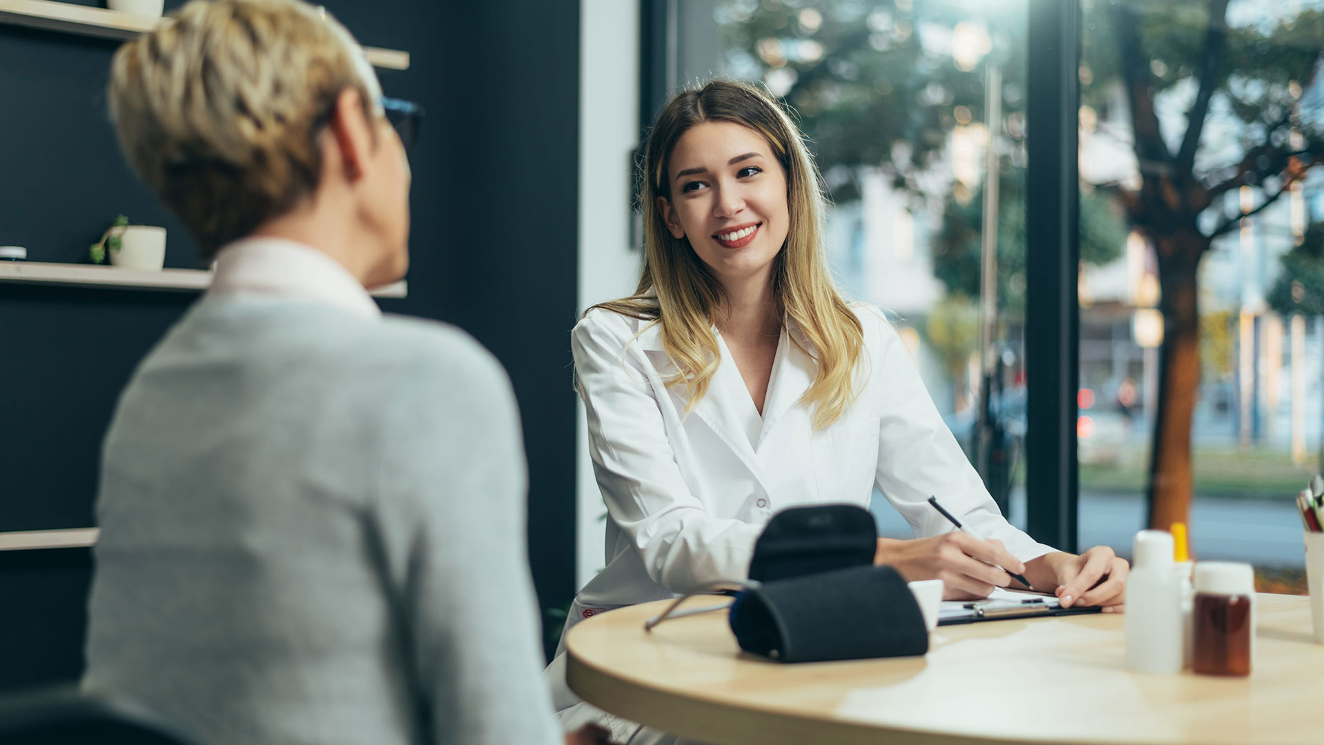 A woman in a white coat smiling while sitting across a table from another person in a consultation setting.