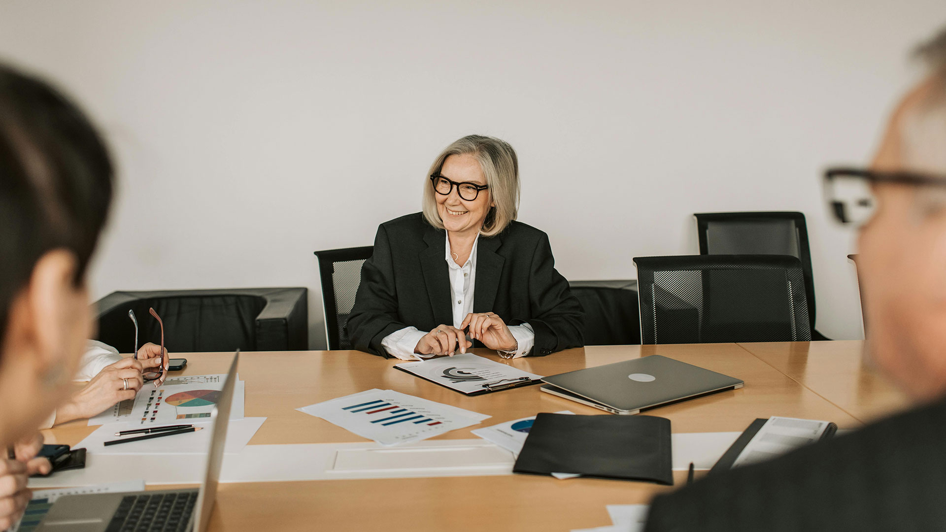 An older woman in business attire smiling at a conference table with colleagues and charts.