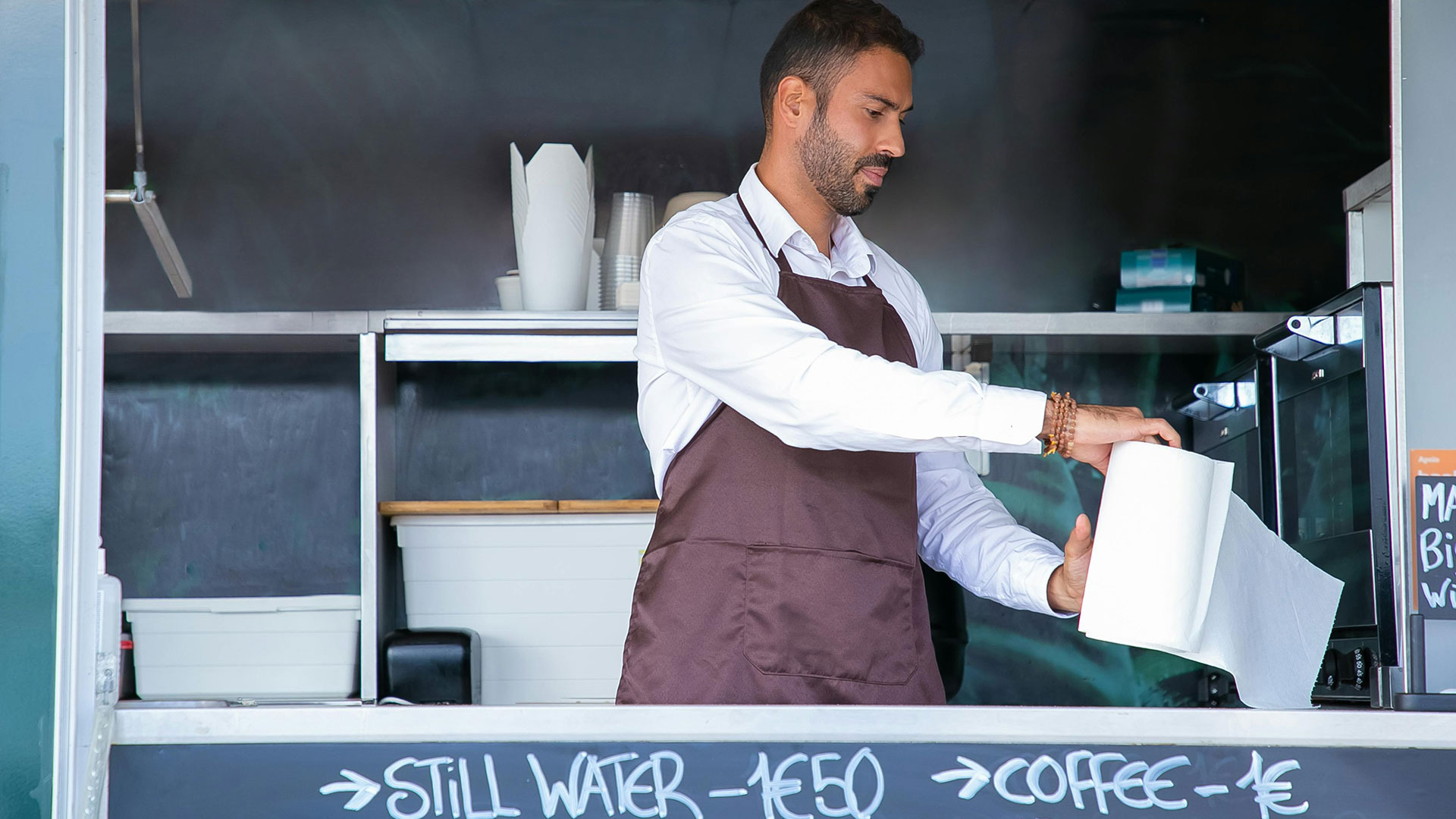 A man in an apron preparing items inside a food truck with a menu chalkboard in front.