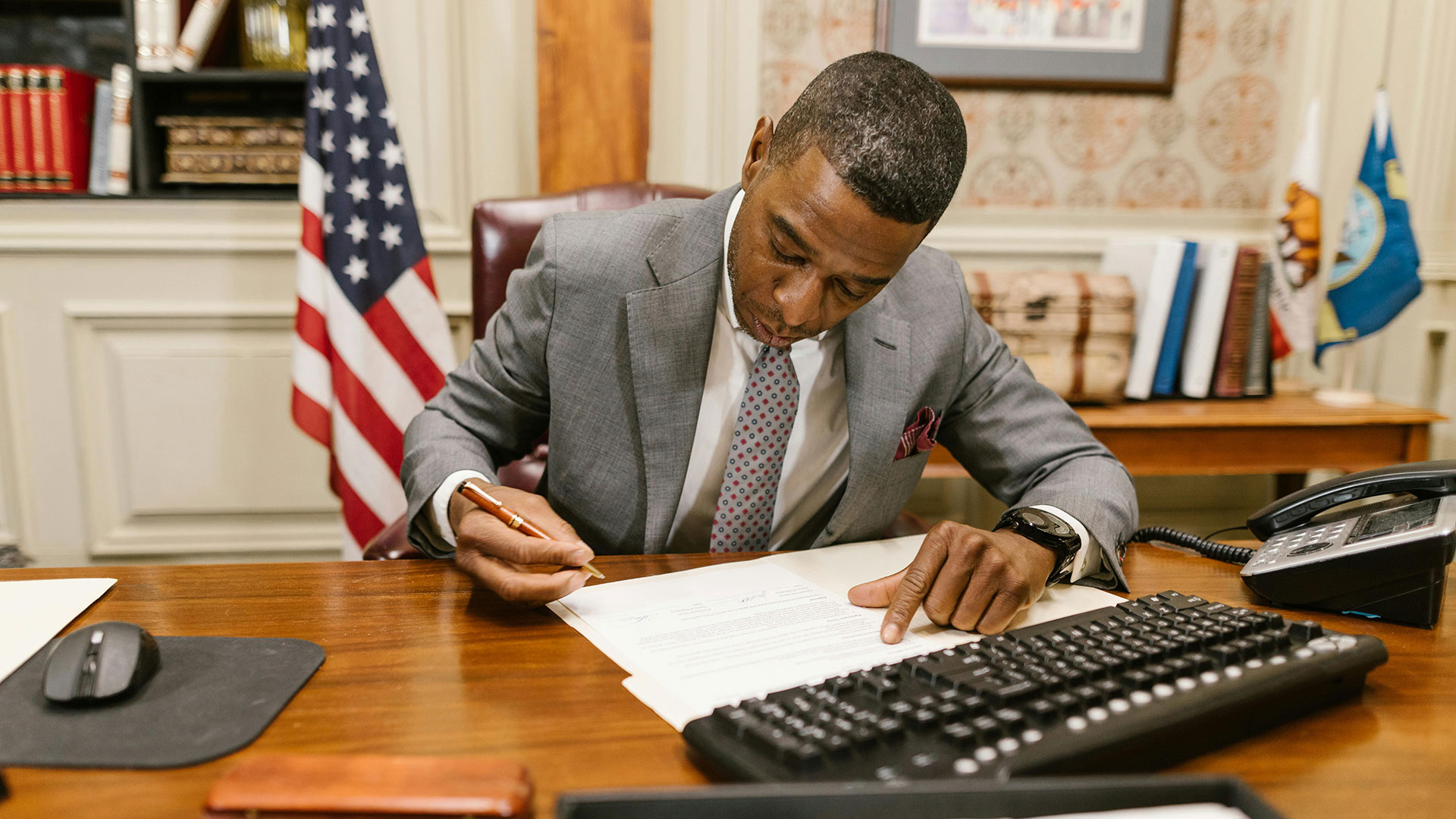 A man in a suit signing documents at a desk with an American flag behind him.