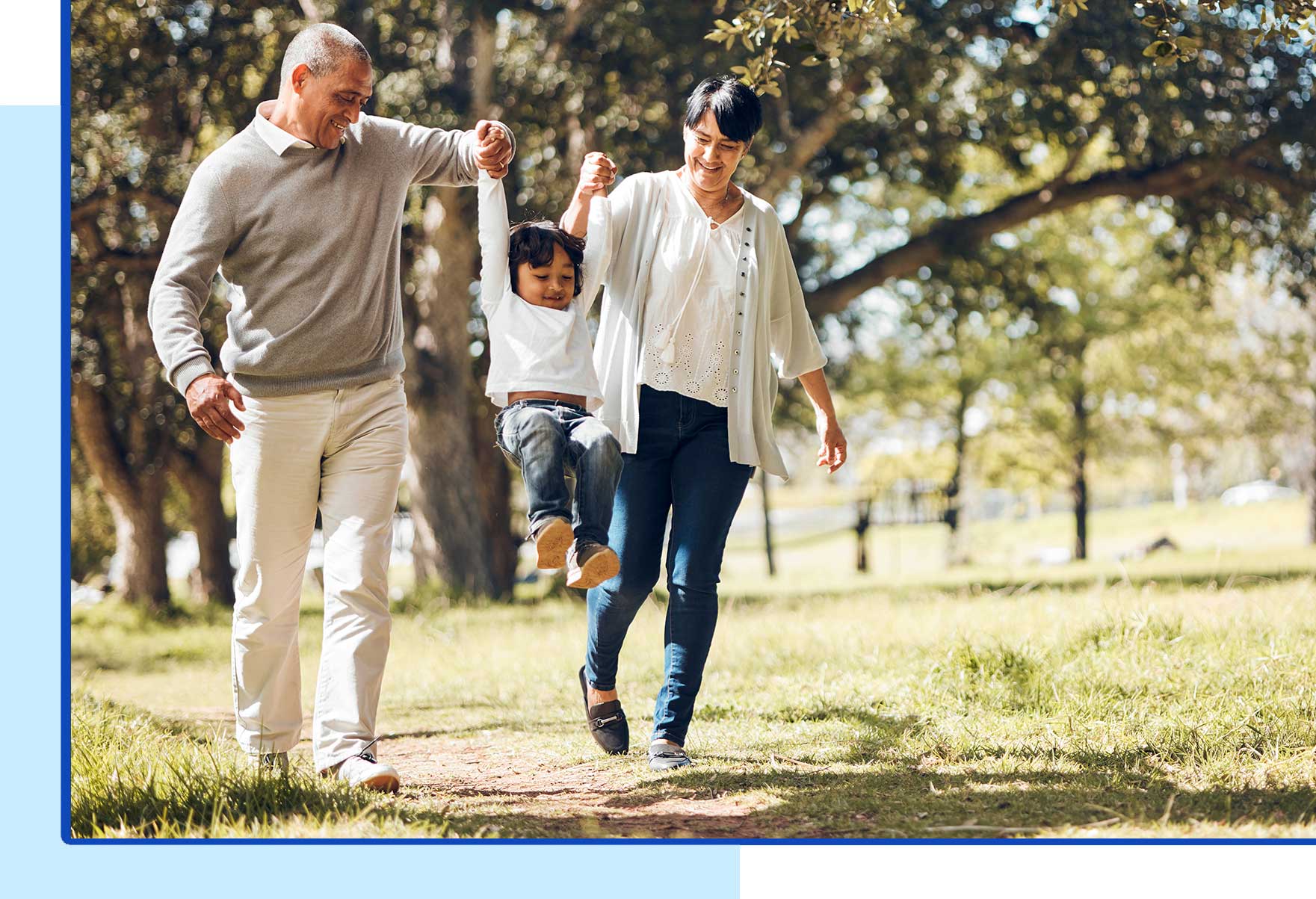 A smiling couple swings a young child by the arms while walking together through a sunny park