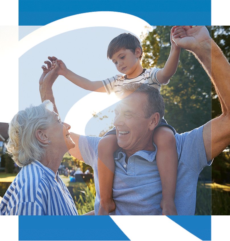 Grandparents smiling outdoors as their grandson sits on his grandfather’s shoulders in the sunlight.