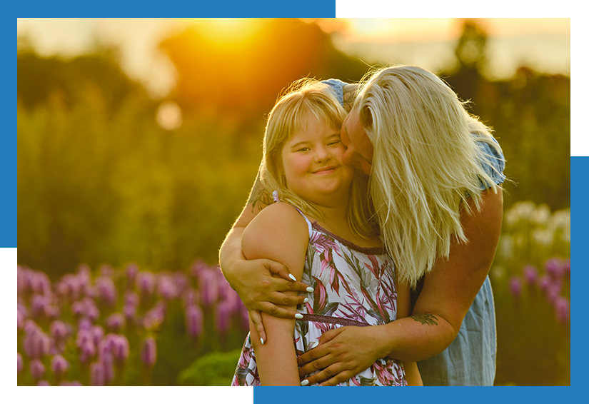 Woman hugging and kissing a smiling girl in a sunlit field of flowers at sunset.