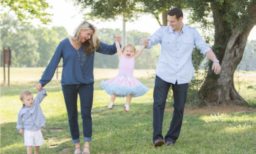 Family walking together swinging daughter outside green trees sunshine