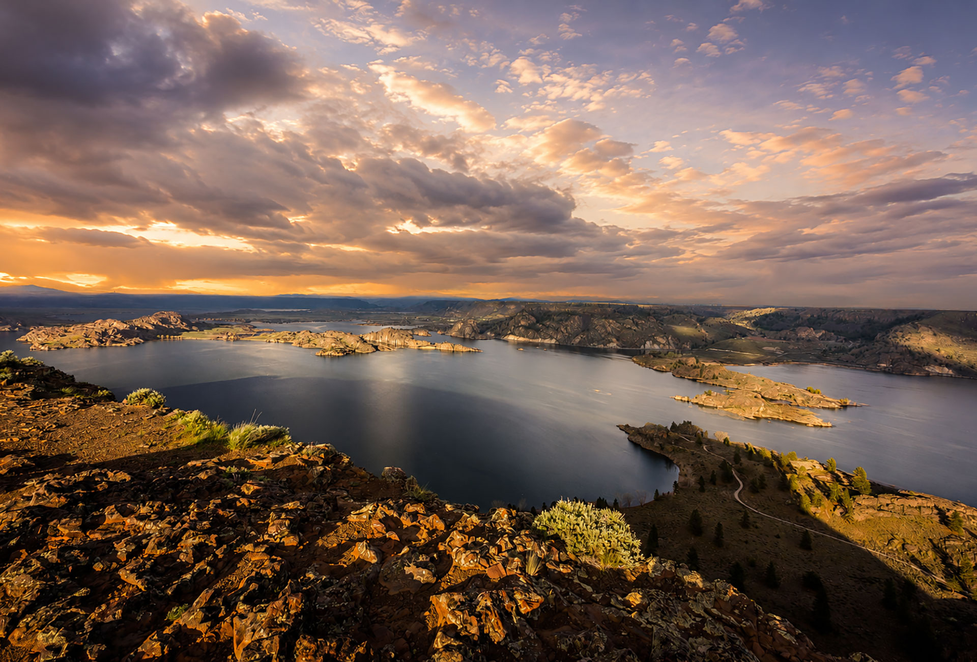 A dramatic sunset paints the sky in orange and gray over a vast lake dotted with rocky islands, viewed from a lichen-covered ridge in the high desert.