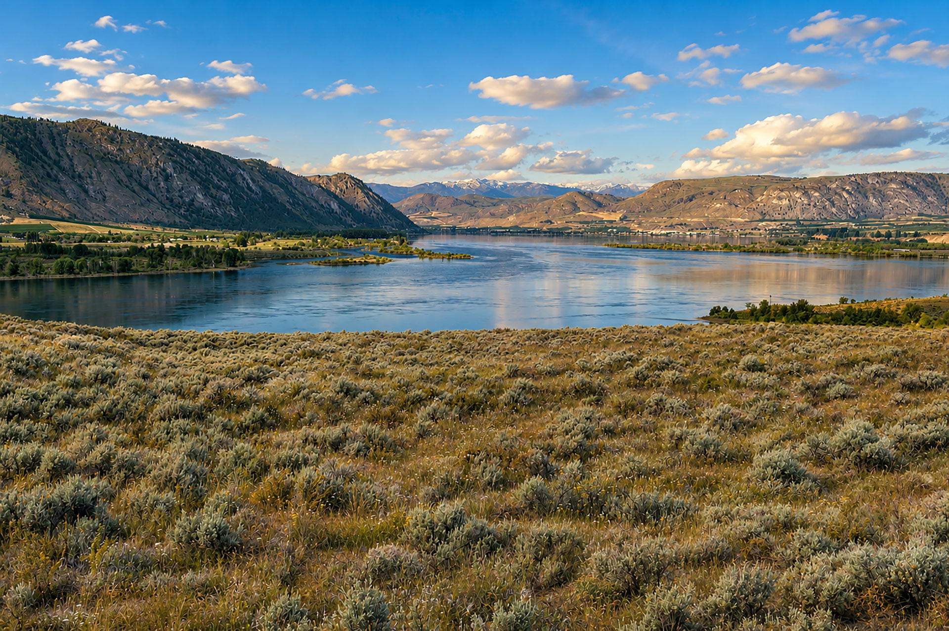 A wide, calm river winds through a valley framed by rolling hills and distant snow-capped peaks, with sagebrush-covered high desert stretching across the foreground under a blue sky dotted with clouds.
