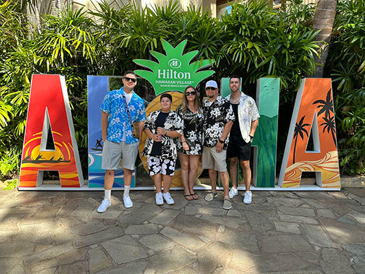 Keri, her husband, and three sons on a trip in front of an "ALOHA" sign