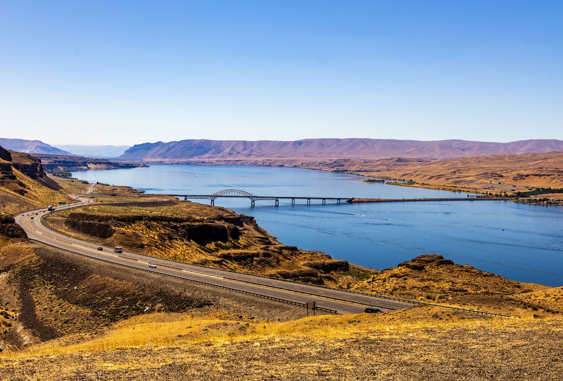 A steel arch bridge and highway curve alongside a wide blue river cutting through golden, arid canyon hills under a clear sky.