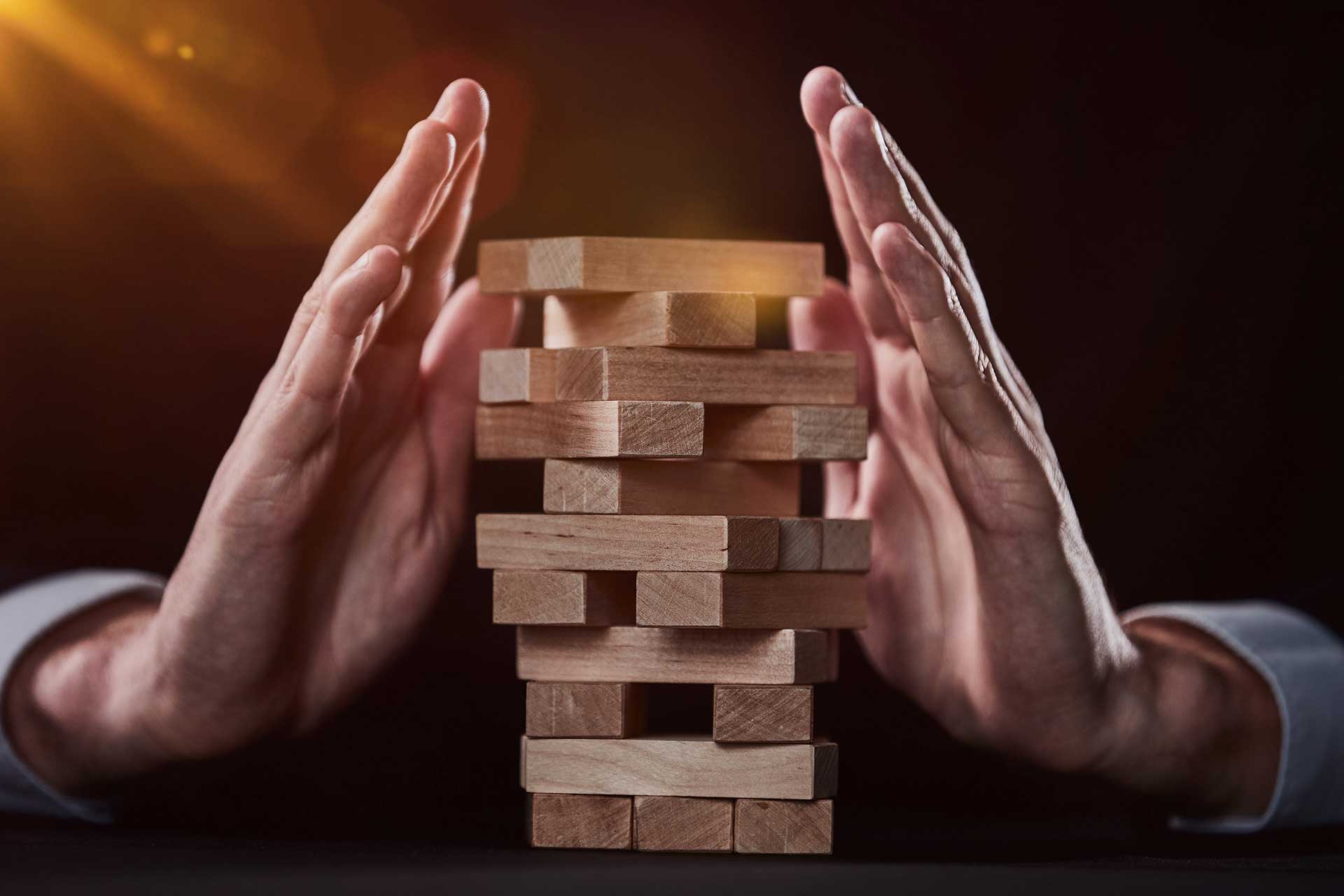 hands hover protectively over a precariously stacked tower of wooden blocks, representing the delicate balance of risk and stability in life and business.