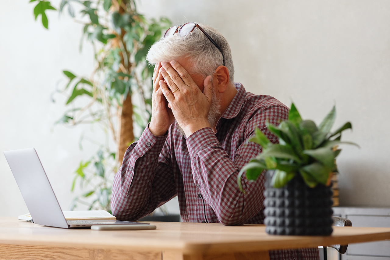 older man at a computer covering his face like he is stressed