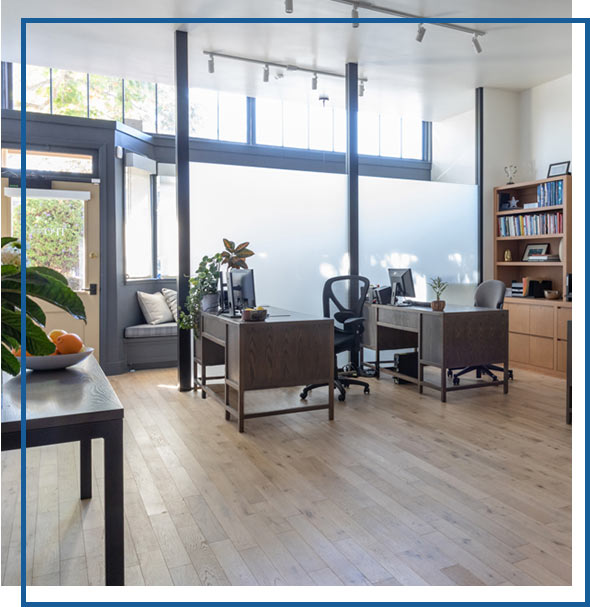 A bright, modern office with large windows, wood floors, and minimalist desks surrounded by plants