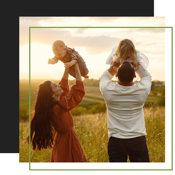 Parents joyfully lifting their young children against a sunset backdrop in a grassy field.