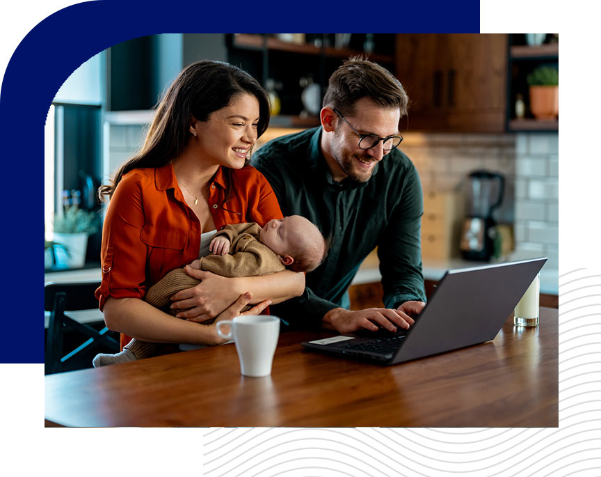 A mom and dad smiling as they sit in the kitchen with coffee, hold their baby, and work on the computer