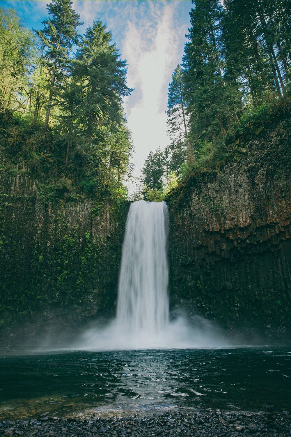 A tall waterfall falls from a forested cliff into a pool below
