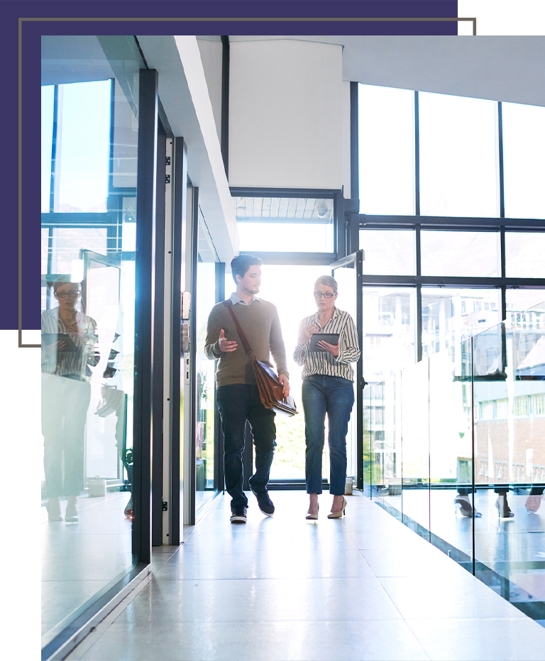 Two colleagues walk through a bright, modern office hallway with floor-to-ceiling windows, engaged in conversation.