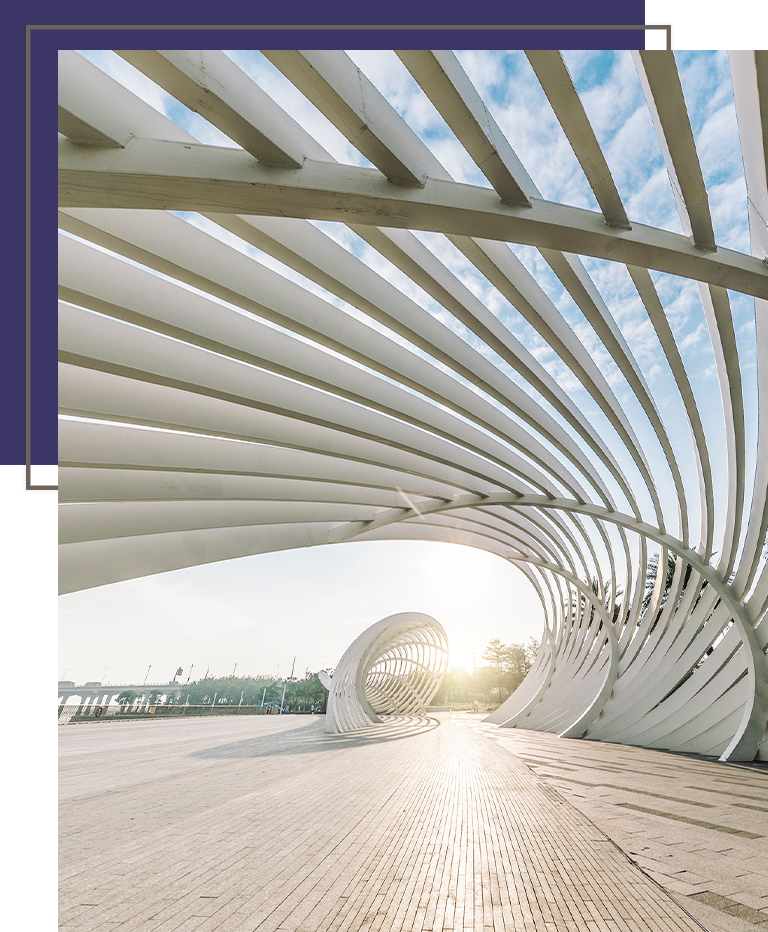 Curved modern architectural structure with white ribs casting shadows on a sunlit walkway beneath a blue sky.