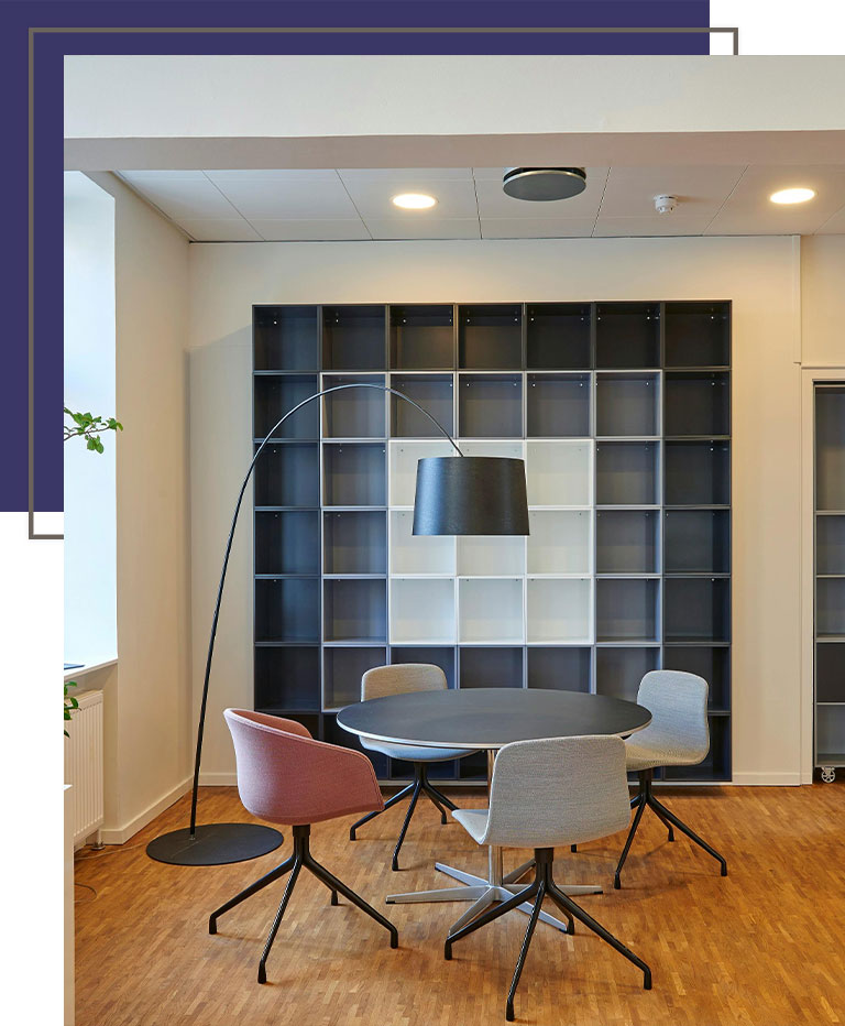 Modern office meeting nook with a round table, colorful chairs, and a tall arc floor lamp set against a grid-style shelving unit.