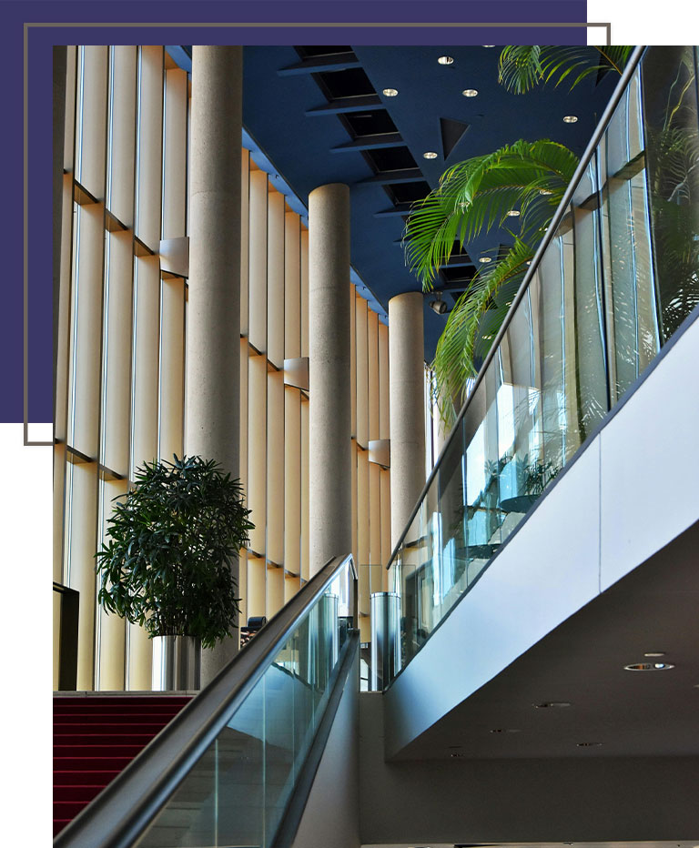 Modern interior with tall columns, a blue ceiling, and lush indoor plants lining a staircase with glass railings.