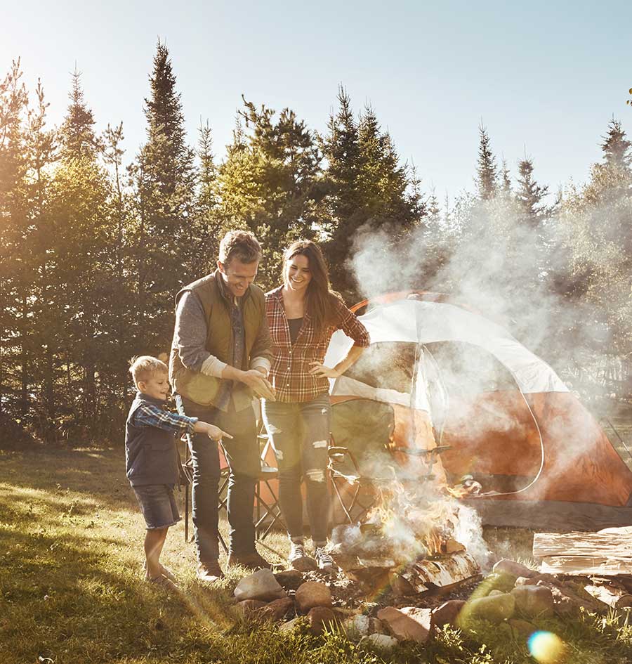 A family enjoying a campsite together, standing by a crackling campfire in front of a tent surrounded by trees.