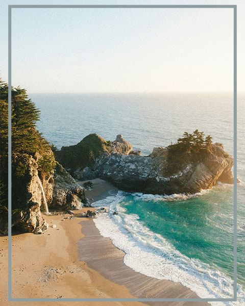 A secluded cove with turquoise waves, a sandy beach, and a waterfall cascading beside rugged coastal cliffs.