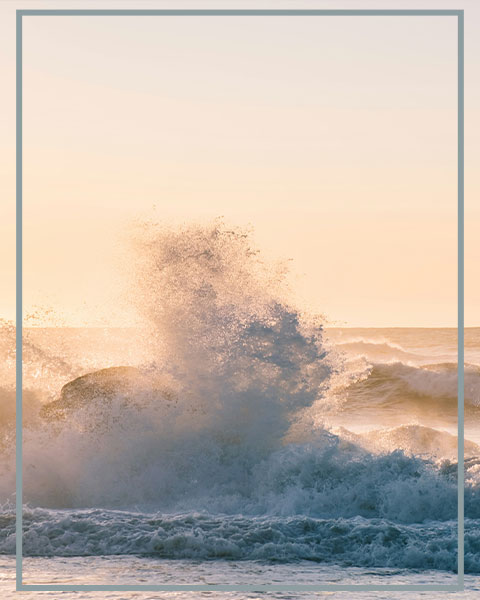 Sunlit ocean waves crash against dark rocks, sending spray into the air at golden hour.