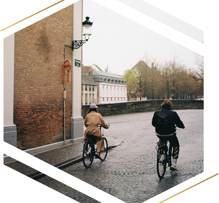 Two people riding bicycles down a cobblestone street in a European town, with brick buildings and bare trees in the distance.