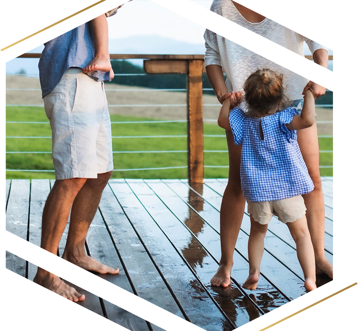 A family of four standing barefoot on a wet wooden deck overlooking a green landscape, with the man holding a small child and the woman holding hands with a toddler.