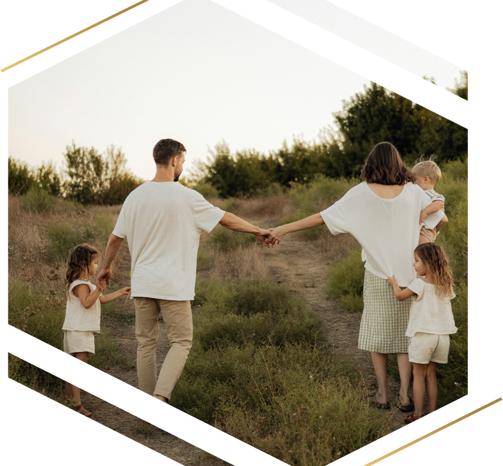 A family of five walking away down a dirt path through a grassy field, with the parents holding hands and each holding a child's hand, and a small child being carried.